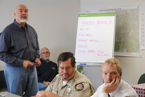 Mike Esnard (standing), president of the Mountain Communities Fire Safe Council, leads the discussion with local fire and recreation leaders about preparing a revised Community Wildfire Protection Plan. Sitting in the background is Chris Kramer of the FSC. Sitting to the right of Esnard are Paul Reisman (center), superintendent of the Mt. San Jacinto State Park, and Idyllwild Fire Chief Patrick Reitz.       Photo by J.P. Crumrine