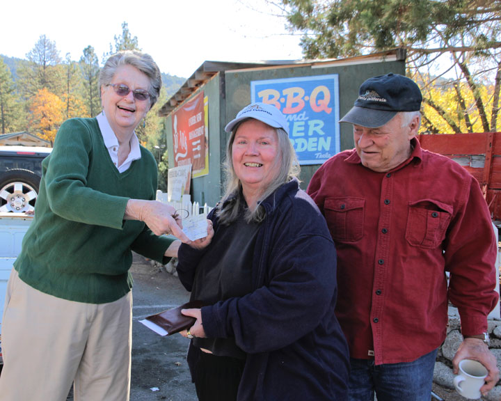 Judy Begin (left) of the Idyllwild Gift Shop, hands to Phyllis Mueller, one of the top dogs of the Tree Lighting Ceremony executive team, a donation to save the Christmas tree that is threatened by the drought, while Joe McNabb, who assisted with mounting the lights, looks on Saturday.Photo by John Drake