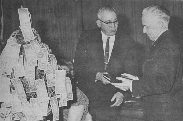 Veteran So. Edison Power Co. worker Glenn Froehlich (right) receives a book of greetings from Walter Lyell in a party in early 1965 in Hemet honouring Froehlich upon his retirement. The money tree hides Nina Mae, his wife, also honored at the event. File photo 