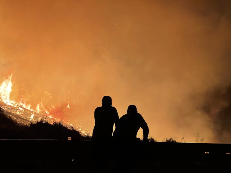 Silhouetted figures observe a large wildfire engulfing the landscape, filling the sky with orange flames and smoke.