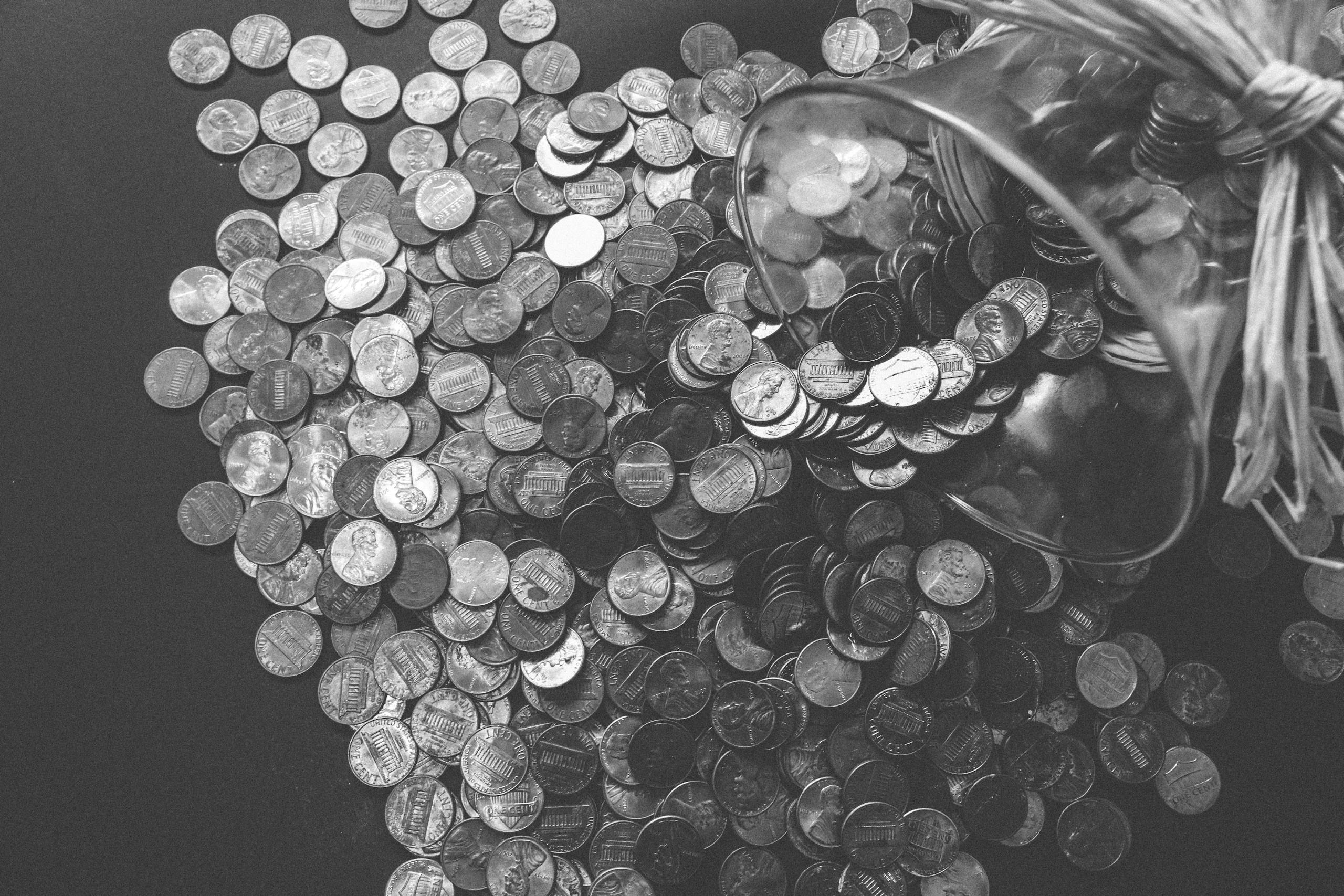 Monochrome image of coins spilling from a glass jar, symbolizing wealth and savings.