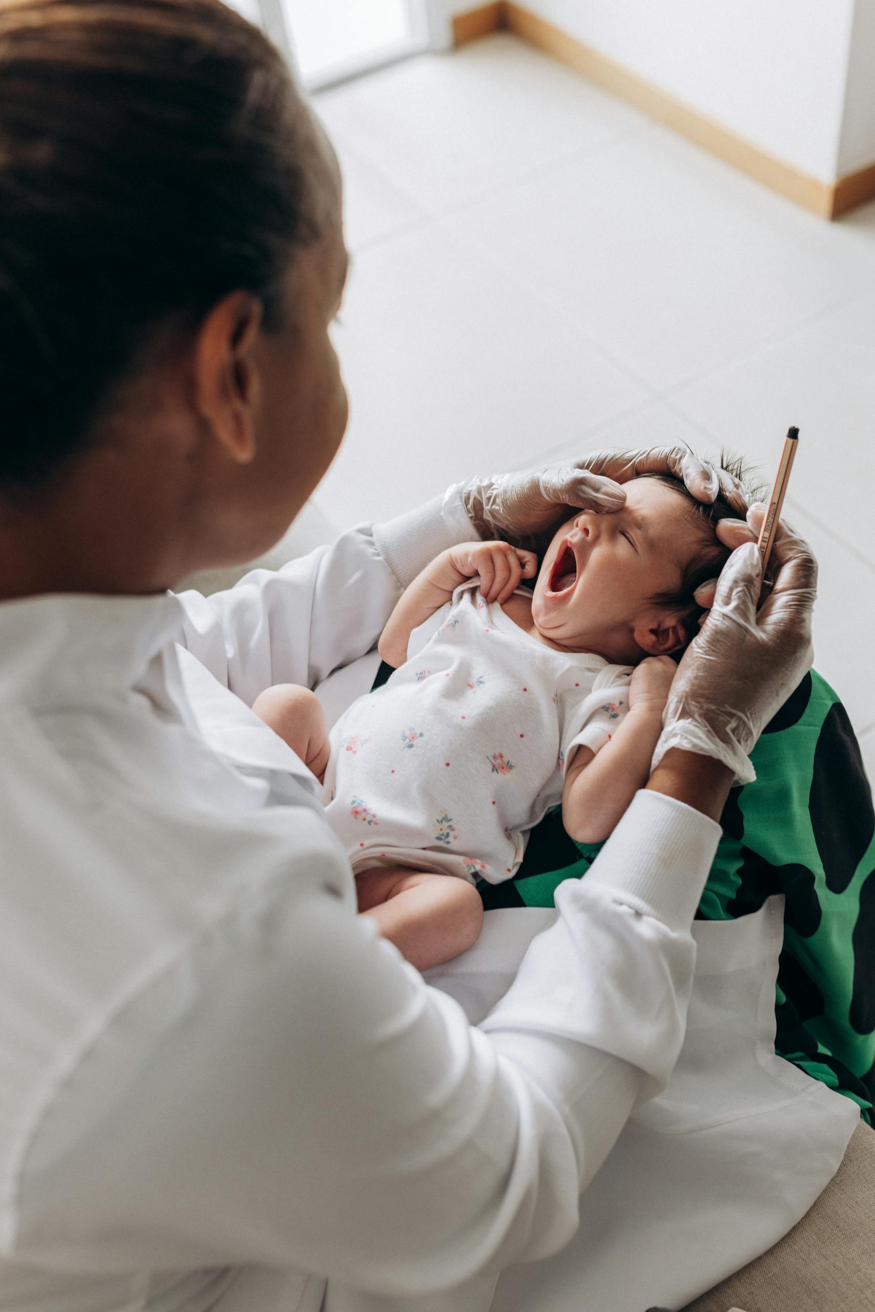 A newborn baby yawns during a health checkup by a doctor in a clinical setting.