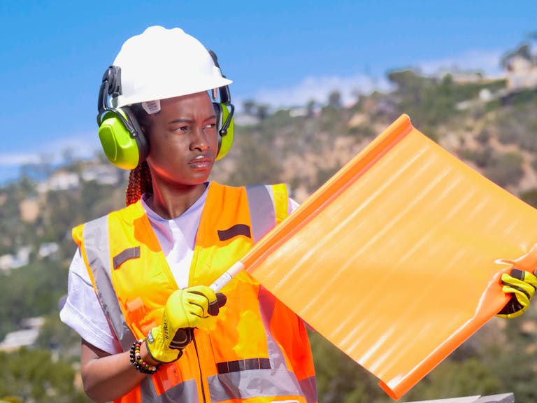 A female flagman wearing a hardhat, earmuffs, and reflective vest holds a safety flag outdoors.