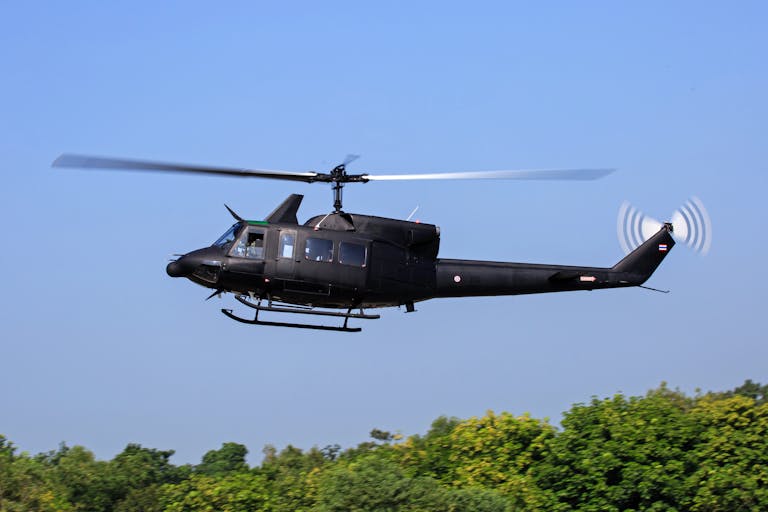 A black military helicopter in flight over lush green trees against a clear blue sky.