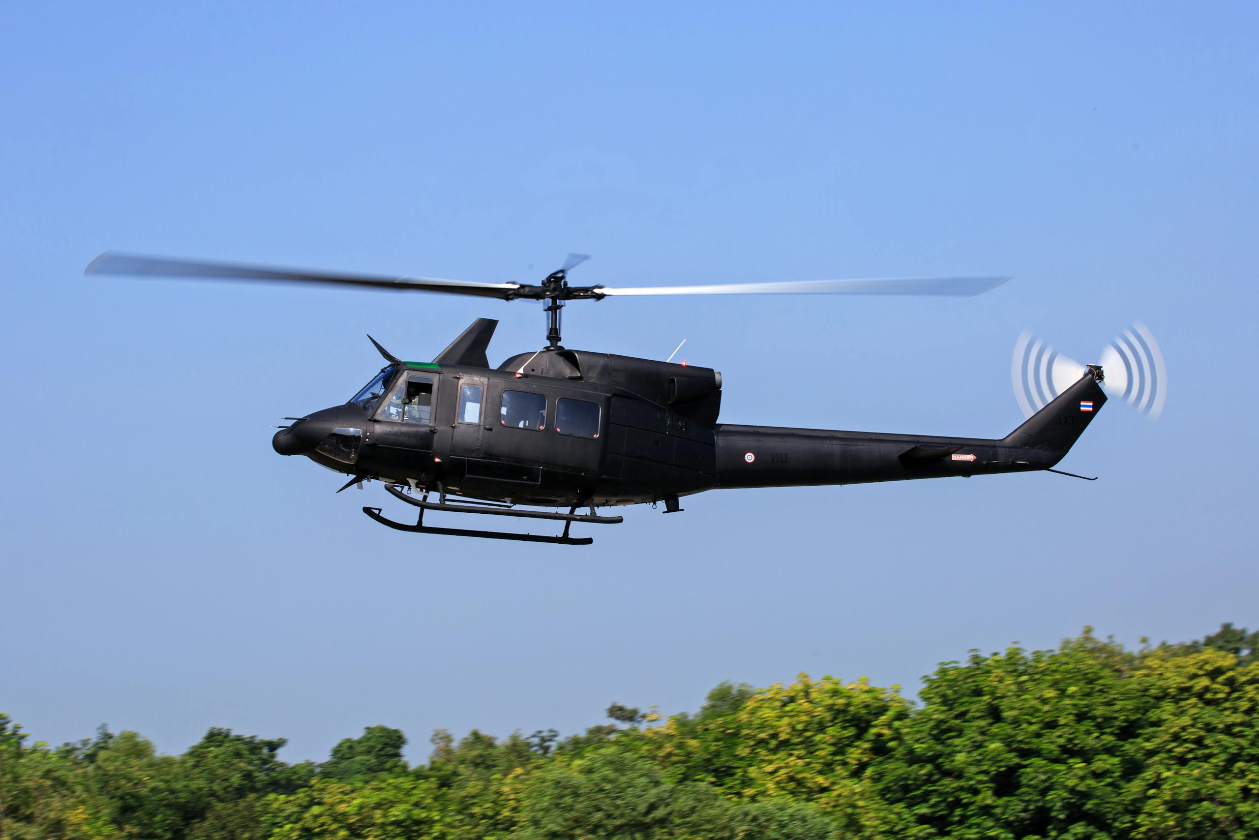 A black military helicopter in flight over lush green trees against a clear blue sky.