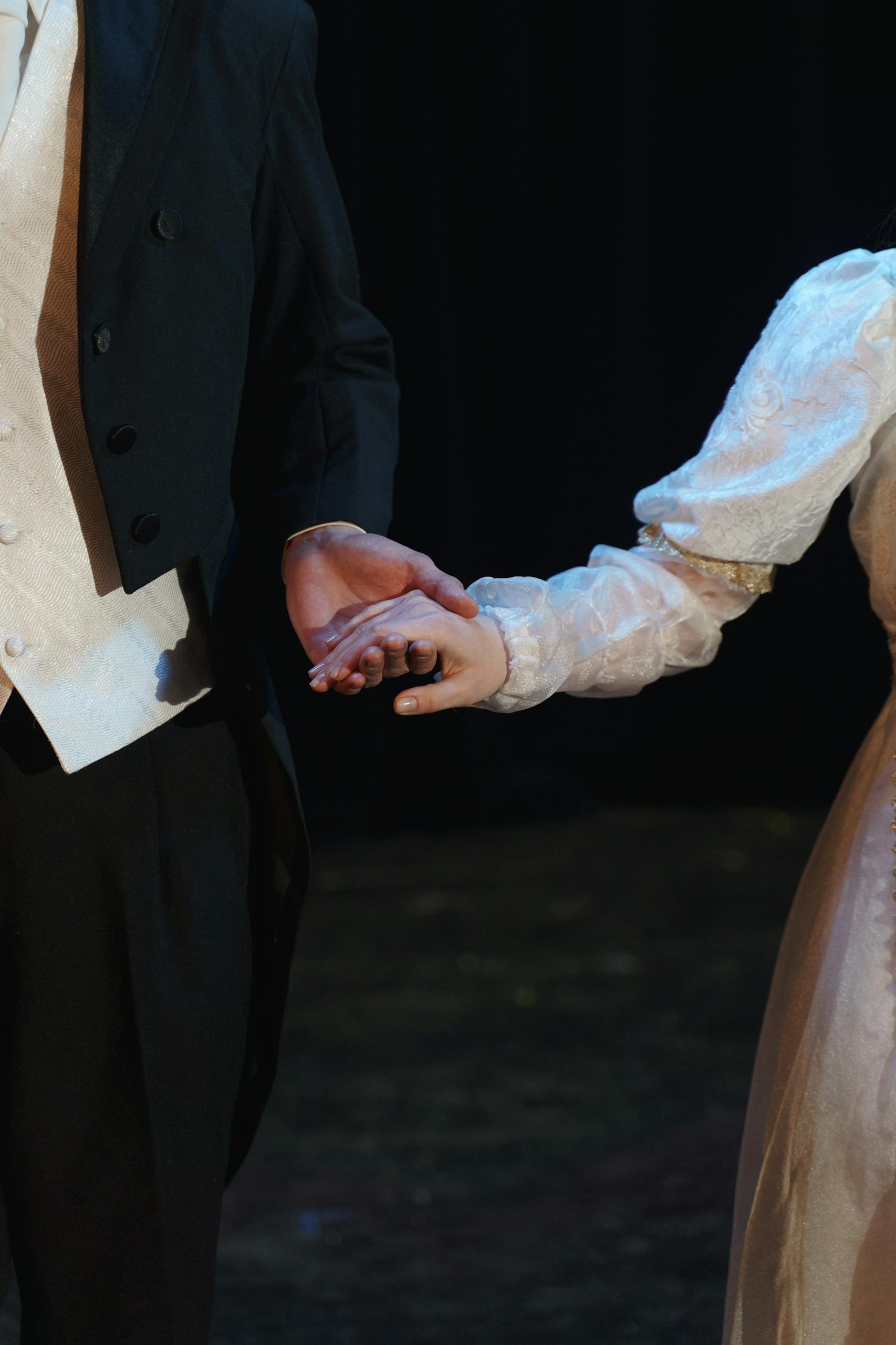 A close-up of two costumed performers holding hands on stage, evoking romance in a theatrical setting.