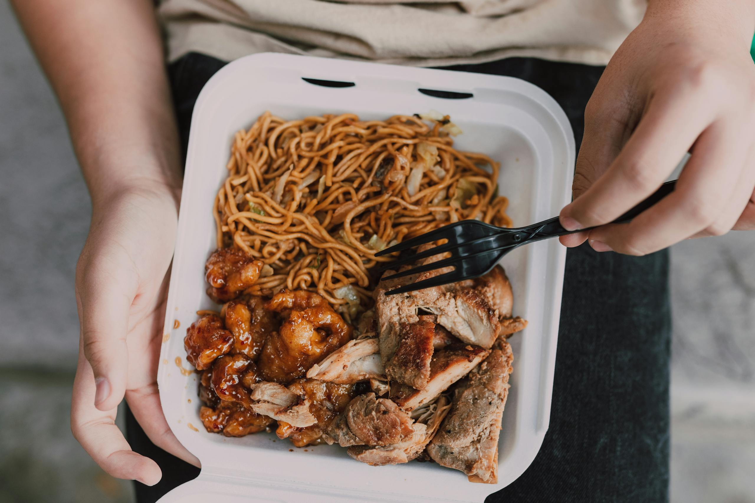 Close-up of takeout box with noodles, chicken, and pork, held in hands.