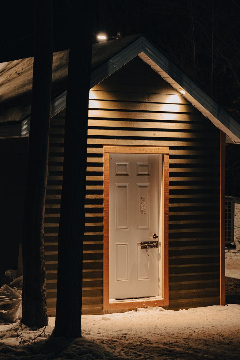 A warmly lit cabin with a white door surrounded by snow, creating a cozy winter night atmosphere.