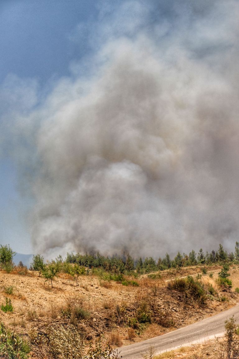 Smoke clouds rise from a forest fire in a rural area with brown grass and dirt roads.