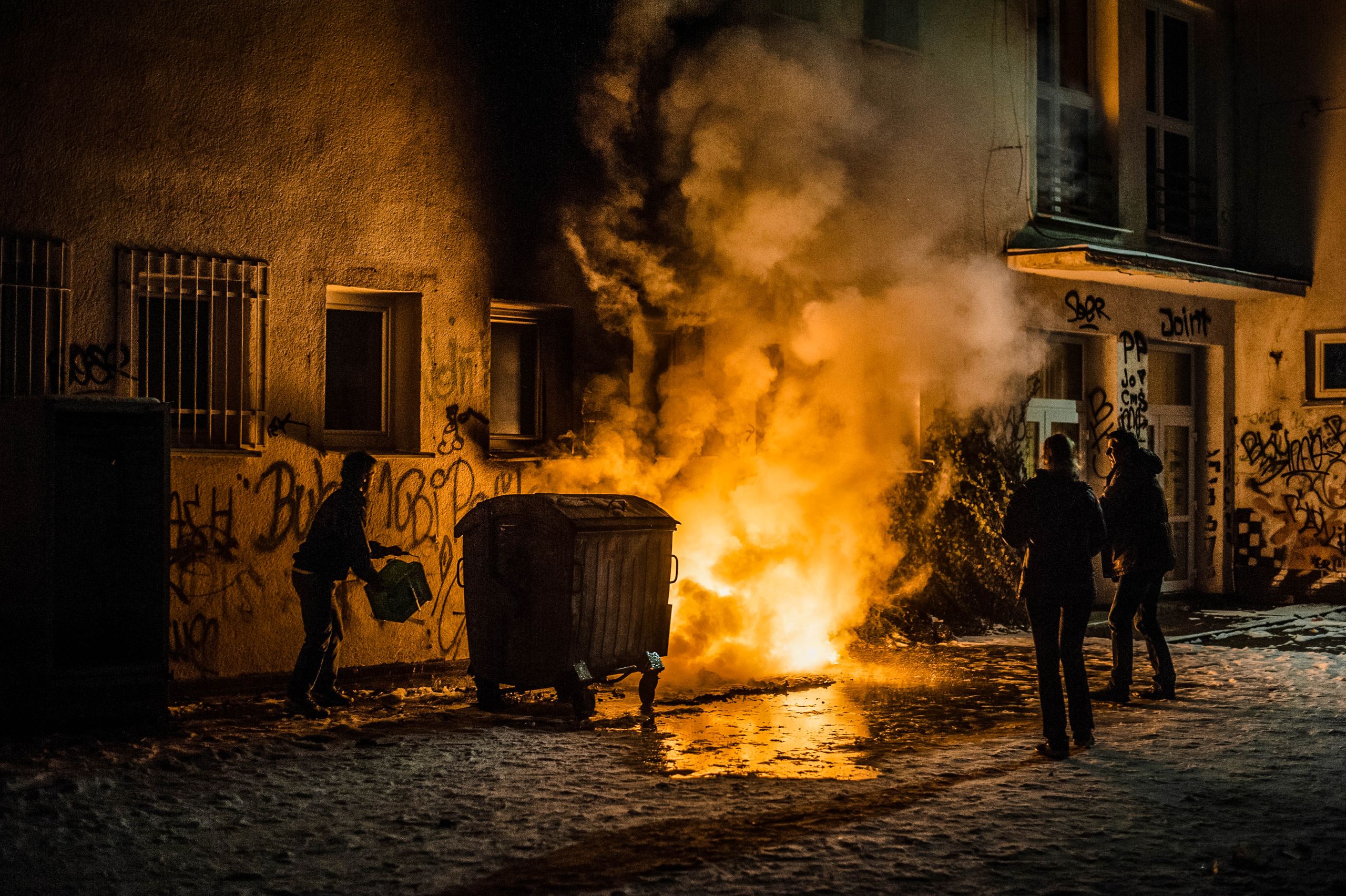 A dramatic urban night scene capturing people near a dumpster fire with graffiti-covered walls.