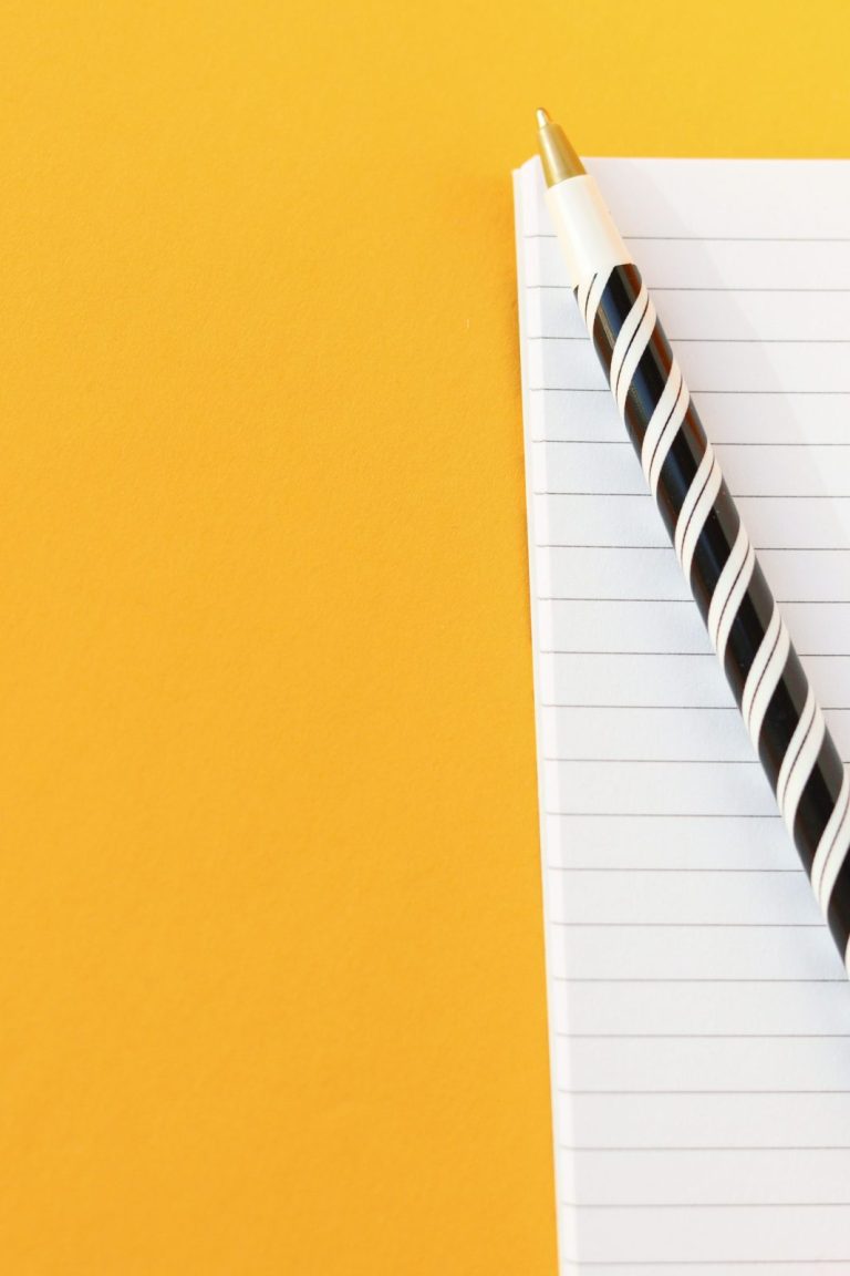 Stock photo for illustration: Black and white striped pen resting on an open lined notebook against a vivid yellow background.